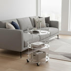 3-tier clear acrylic rolling cart styled in a bright living room, with a vase and decor on top, books on the middle shelf, and a storage basket on the bottom