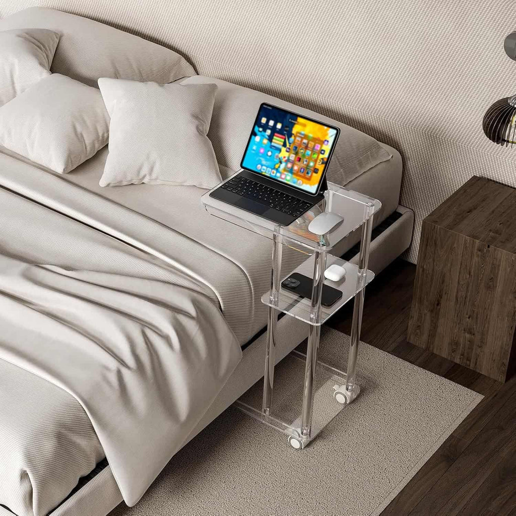 A 3-tier acrylic rolling side table used as a bedside table, holding a laptop, mouse, and phone