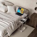 A 3-tier acrylic rolling side table used as a bedside table, holding a laptop, mouse, and phone