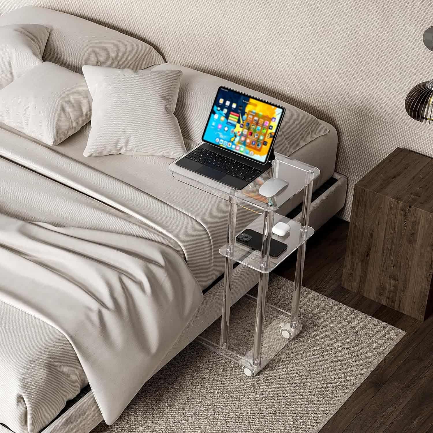 A 3-tier acrylic rolling side table used as a bedside table, holding a laptop, mouse, and phone