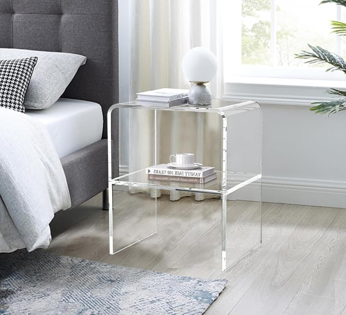 Clear acrylic side table used as a nightstand by the bed, paired with a table lamp and books in a modern bedroom