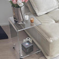 Clear acrylic C-shaped side table styled next to a beige sofa, holding a vase of flowers and candles.