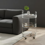 Minimalist 3-tier acrylic bar cart next to a dark gray sofa, decorated with a leafy plant and books, showcasing its sleek, transparent design